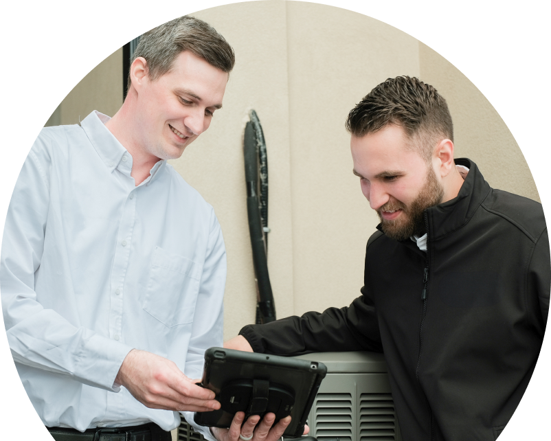 A Anchor Home Comfort technician in a white shirt shows service details on a tablet to a smiling client in a black jacket, standing beside an AC unit outside a beige building. A Anchor Home Comfort technician in a white shirt shows service details on a tablet to a smiling client in a black jacket, standing beside an AC unit outside a beige building.