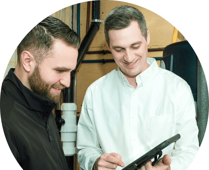 Anchor Home Comfort specialist reviewing maintenance plan options with a smiling homeowner using a tablet, inside a utility room with visible piping. Anchor Home Comfort specialist reviewing maintenance plan options with a smiling homeowner using a tablet, inside a utility room with visible piping.