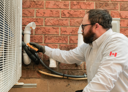 A Anchor Home Comfort HVAC specialist wearing glasses and gloves uses a diagnostic tool to service an outdoor AC unit beside a brick wall. A Anchor Home Comfort HVAC specialist wearing glasses and gloves uses a diagnostic tool to service an outdoor AC unit beside a brick wall.
